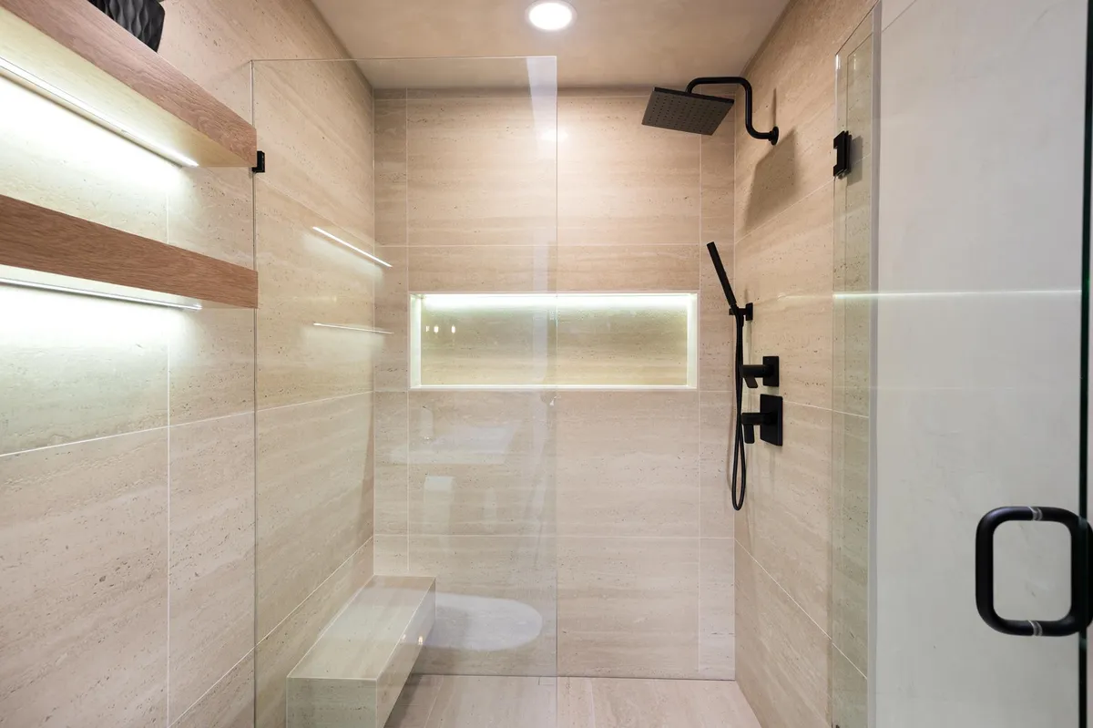 Walk-in shower in travertine-look tile with a lit niche and matte black fixtures